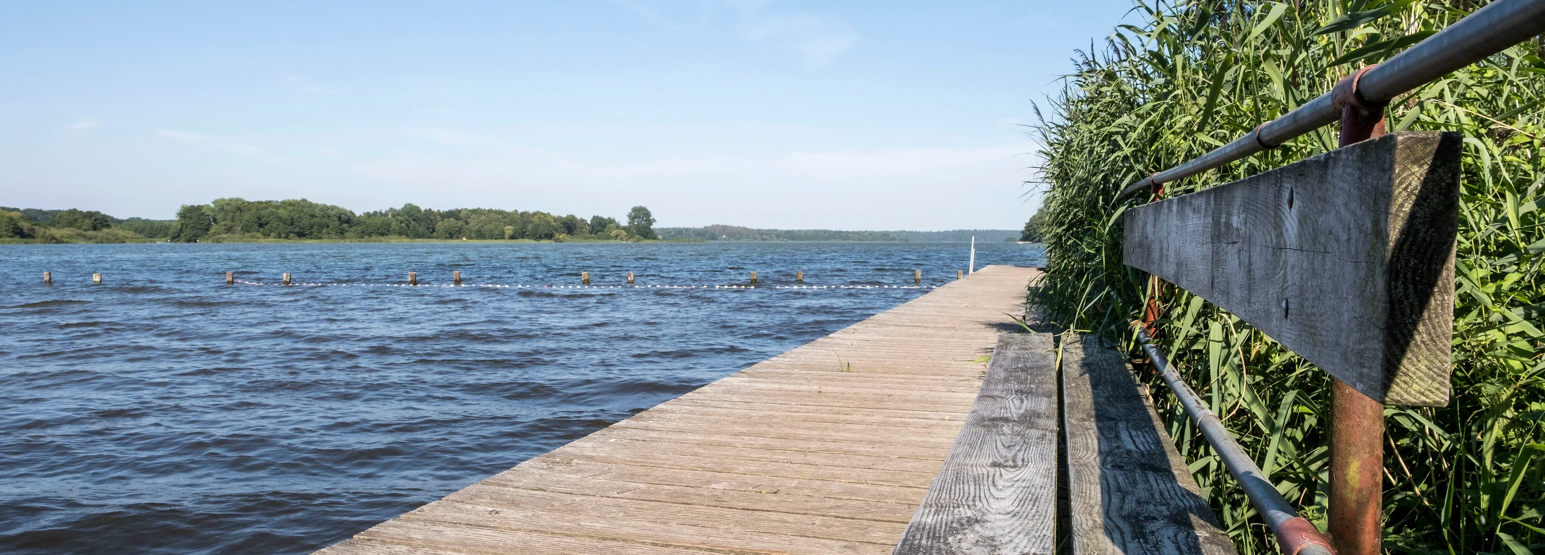 Ein Holzsteg mit einer Holzbank führt in das blaue Seewasser hinein. Der Himmel ist blau und es ist Sommer.