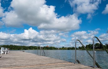 Badestellen & Bäder (© MHT.SH & Peter Lühr) Ein Holzsteg führt in den Bordesholmer See im Hintergrund sieht man eine Kirche. Der Himmel ist blau mit Wolken.