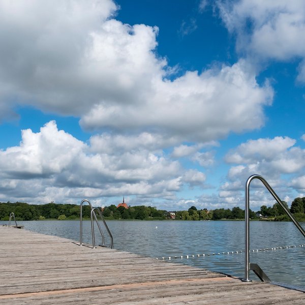 Badestellen & Bäder (© MHT.SH & Peter Lühr) Ein Holzsteg führt in den Bordesholmer See im Hintergrund sieht man eine Kirche. Der Himmel ist blau mit Wolken.