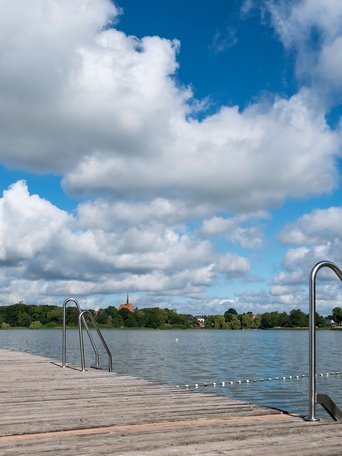 Ein Holzsteg führt in den Bordesholmer See im Hintergrund sieht man eine Kirche. Der Himmel ist blau mit Wolken.