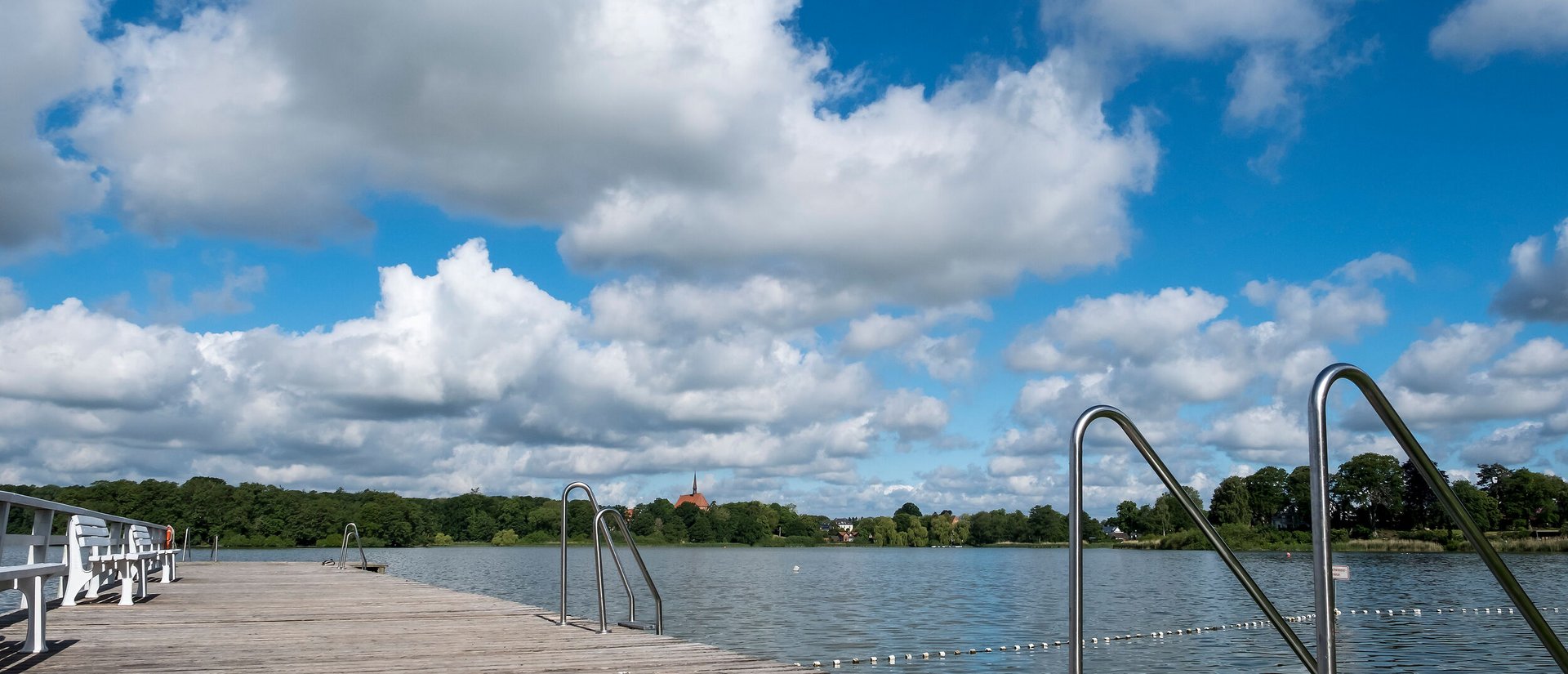 Ein Holzsteg führt in den Bordesholmer See im Hintergrund sieht man eine Kirche. Der Himmel ist blau mit Wolken.