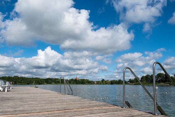 Ein Holzsteg führt in den Bordesholmer See im Hintergrund sieht man eine Kirche. Der Himmel ist blau mit Wolken.