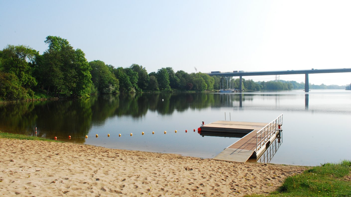 Ein Badesteg am See mit einer Sandfläche. Im Hintergrund ist eine Brücke zu sehen.