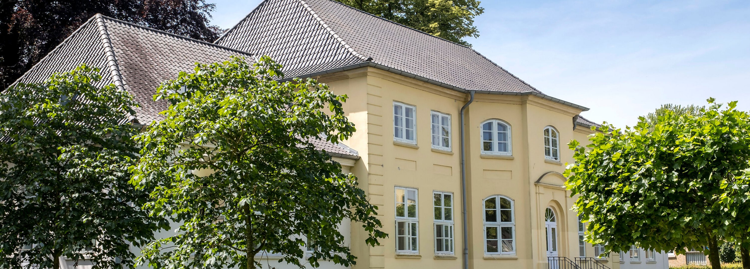 Ein herrschaftliches gelbes Haus umgeben von grünen Bäumen. Der Himmel ist blau, die Sonne scheint.