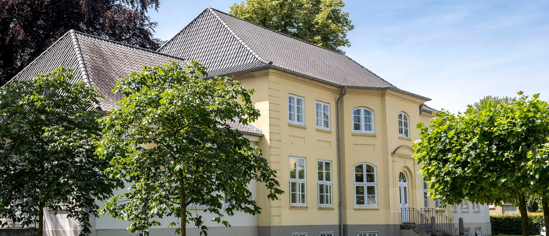 Ein herrschaftliches gelbes Haus umgeben von grünen Bäumen. Der Himmel ist blau, die Sonne scheint.