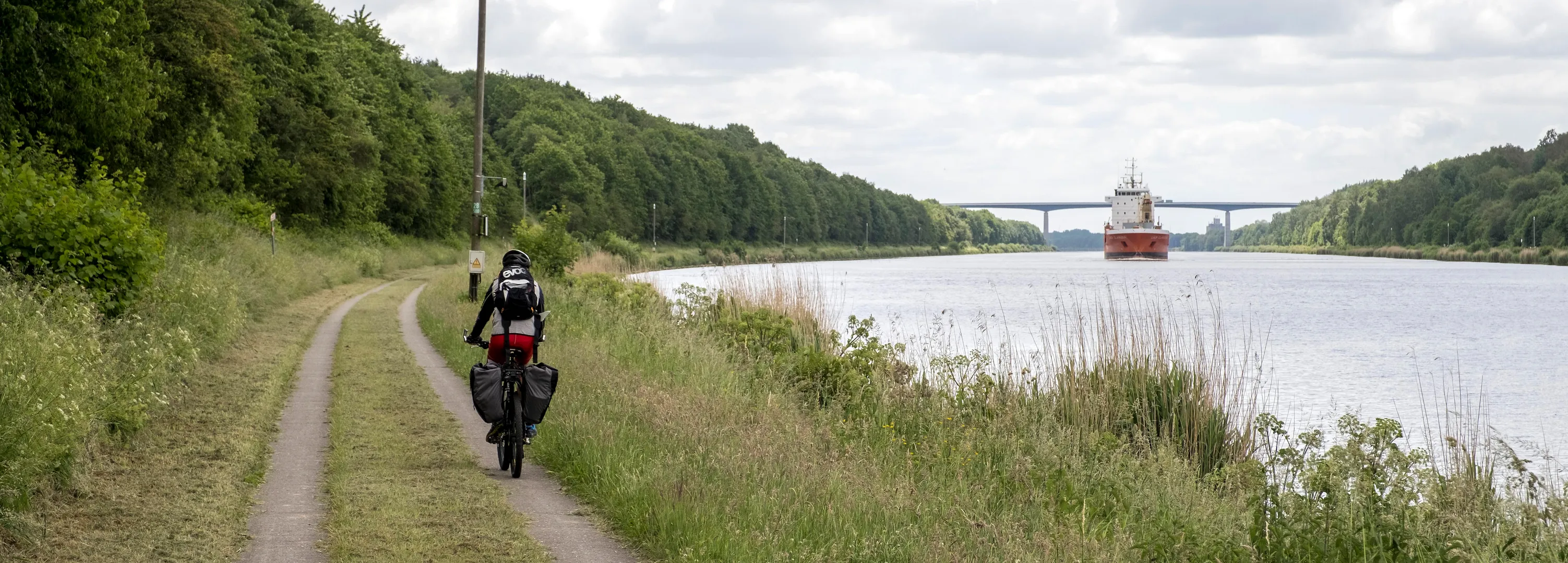 Ein Radfahrer fährt am NOK entlang im Hintergrund sieht man ein Schiff auf dem NOK