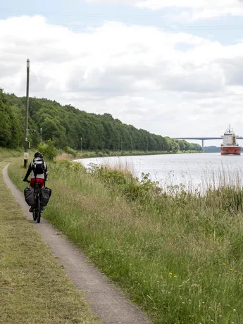 Ein Radfahrer fährt am NOK entlang im Hintergrund sieht man ein Schiff auf dem NOK