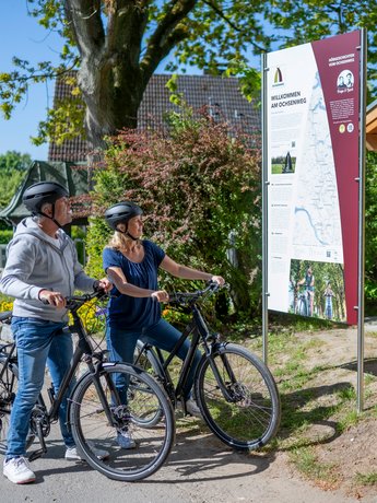 Zwei Menschen stehen mit Rädern vor einem großen Info-Schild. 