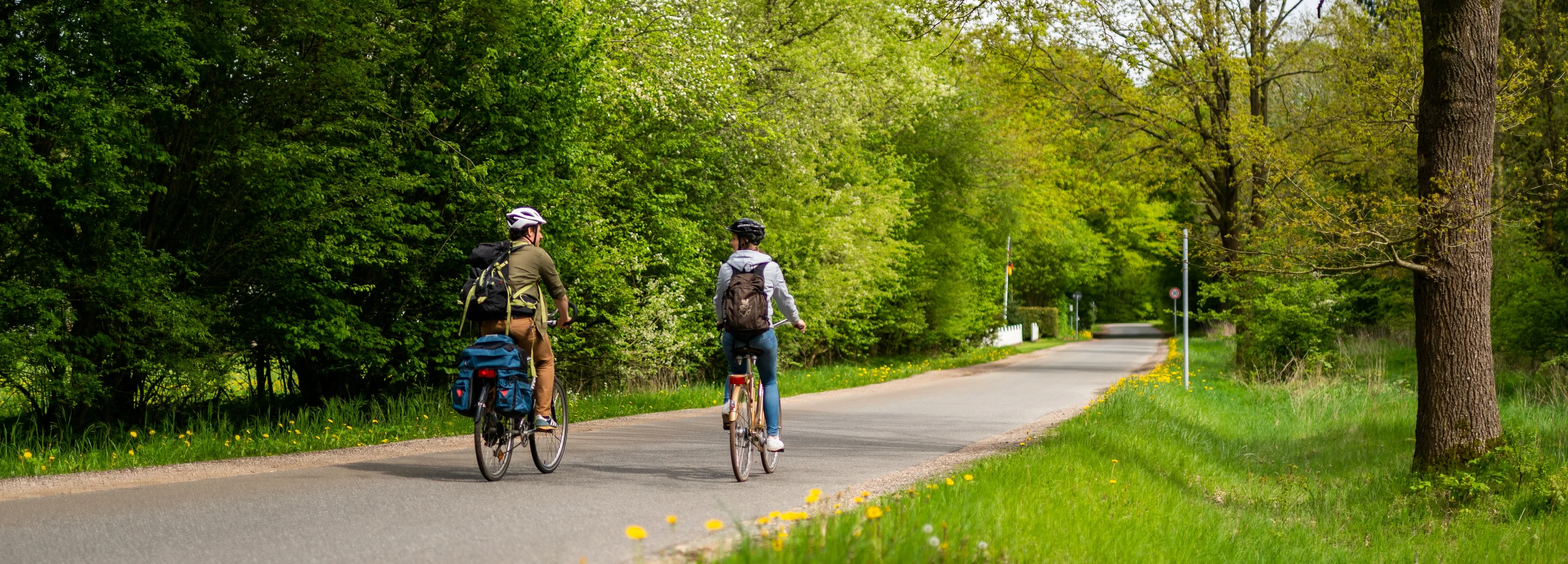 Zwei Menschen fahren auf ihren Rädern durch eine grüne Landschaft. Bäume säume den asphaltierten Weg.