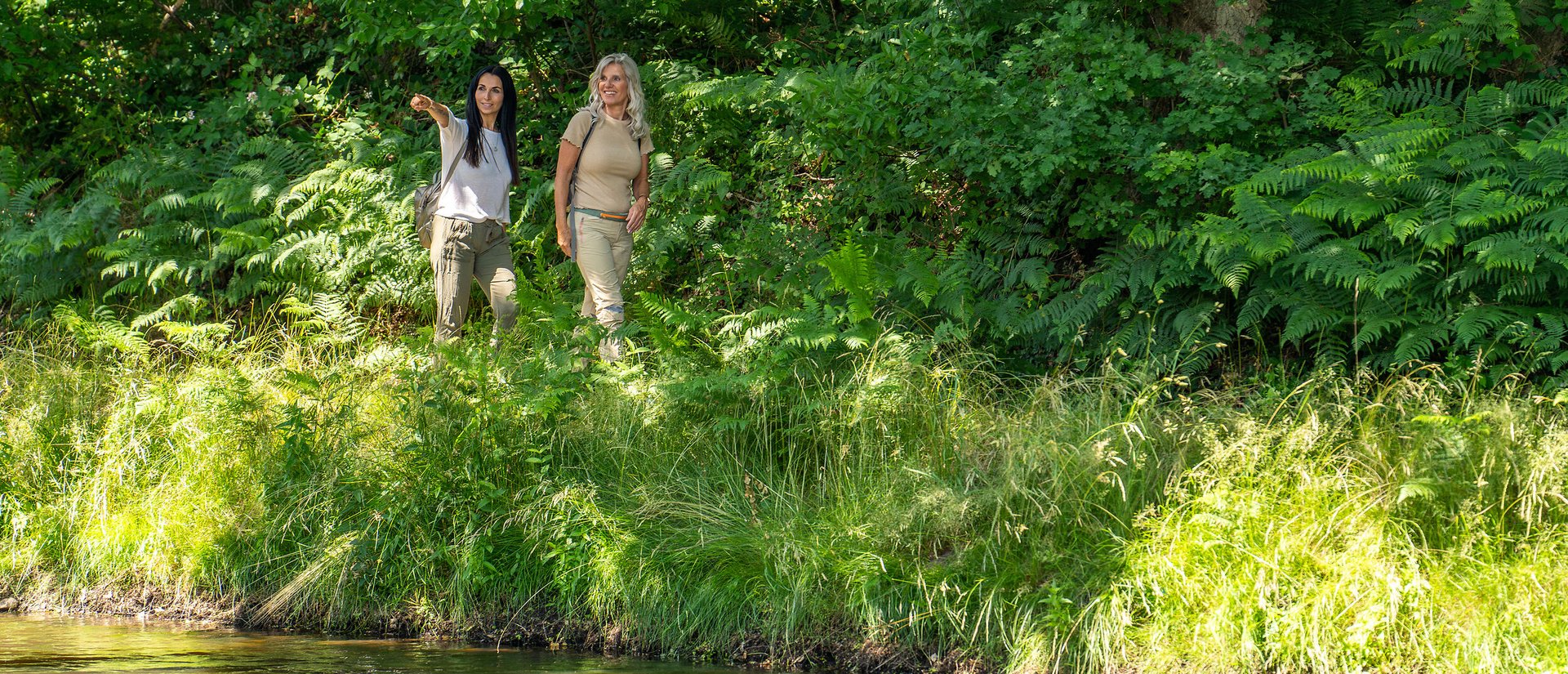 Zwei Frauen wandern durch grüne Landschaft entlang eines Gewässers. 