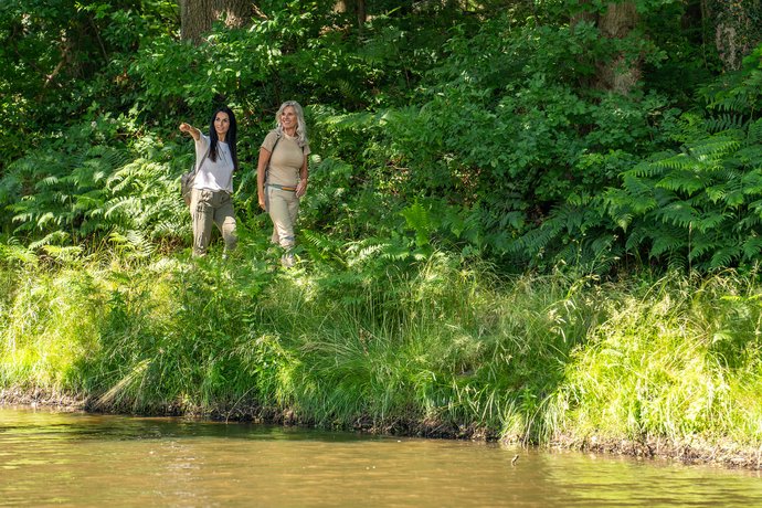 Zwei Frauen wandern durch grüne Landschaft entlang eines Gewässers. 