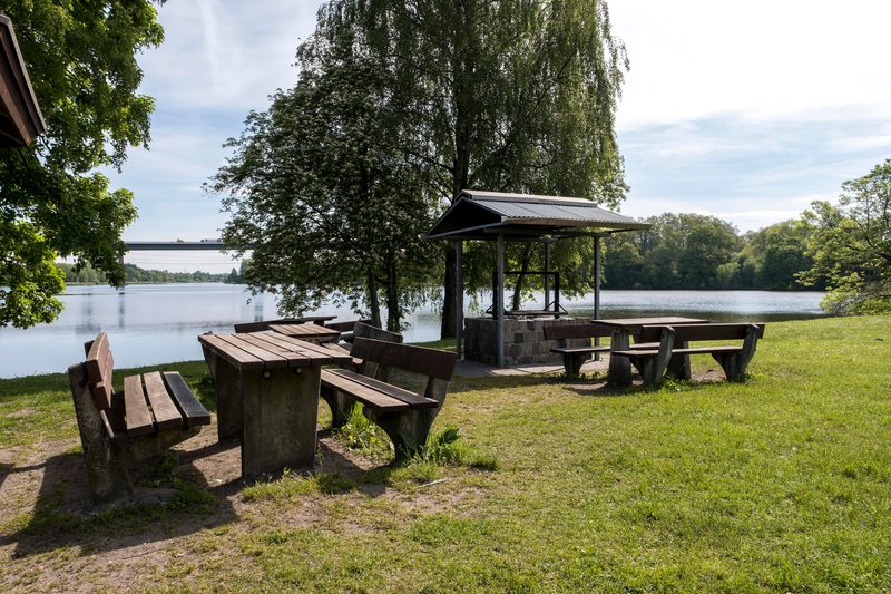 Picknickplatz mit Holzbänken und Tischen am Ufer des Borgstedter Sees, umgeben von Bäumen und mit Blick auf eine Brücke im Hintergrund.