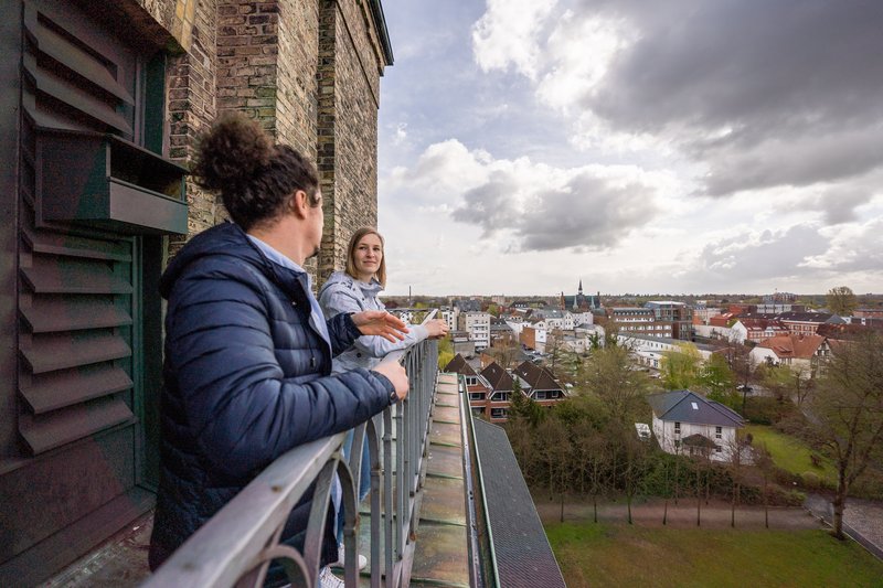 Zwei Personen stehen auf einem Balkon mit Blick auf eine Stadtlandschaft.