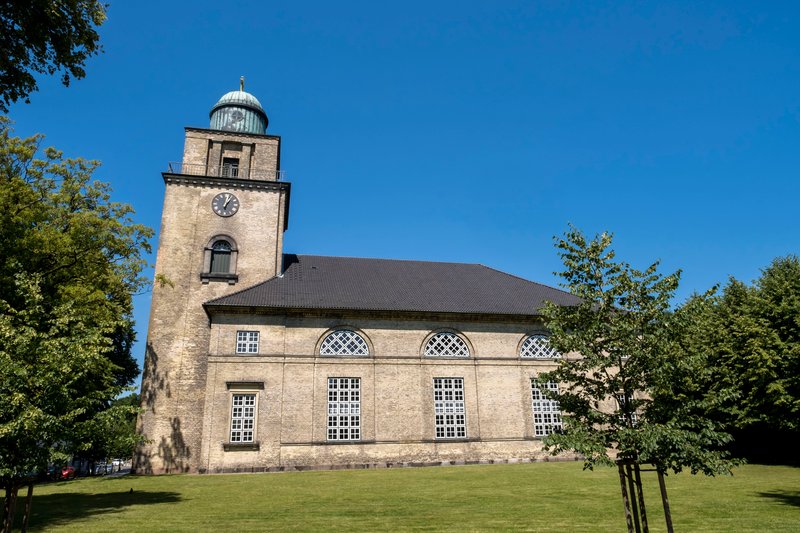 Seitenansicht der Vicelinkirche in Neumünster mit Turm und Uhr, umgeben von Bäumen und Rasenfläche, unter klarem blauem Himmel.