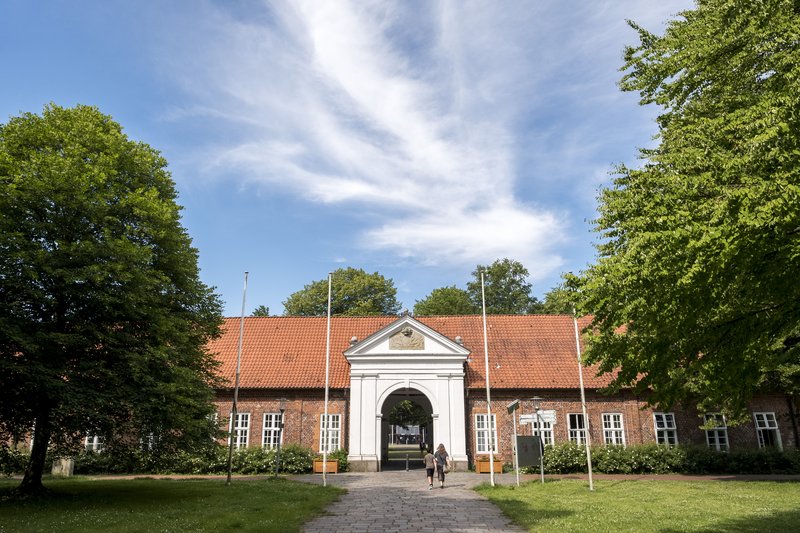 Historisches Gebäude mit rotem Ziegeldach und weißem Torbogen, umgeben von Bäumen und blauem Himmel.