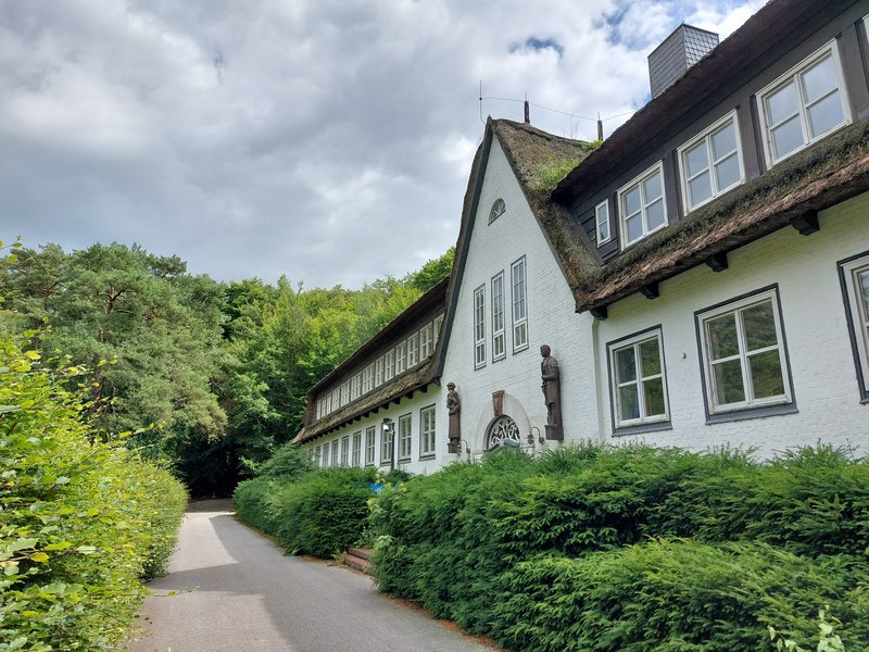 Ein weißes Gebäude mit Reetdach und Statuen an der Fassade, umgeben von grünen Büschen und Bäumen, unter einem bewölkten Himmel.