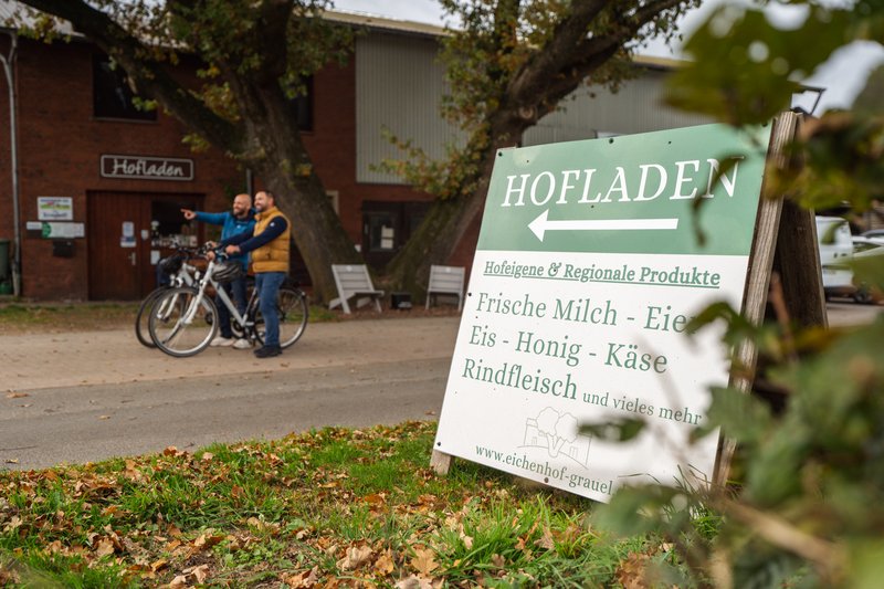 Ein Schild vor einem Hofladen, das auf frische und regionale Produkte hinweist. Im Hintergrund stehen zwei Personen mit Fahrrädern.