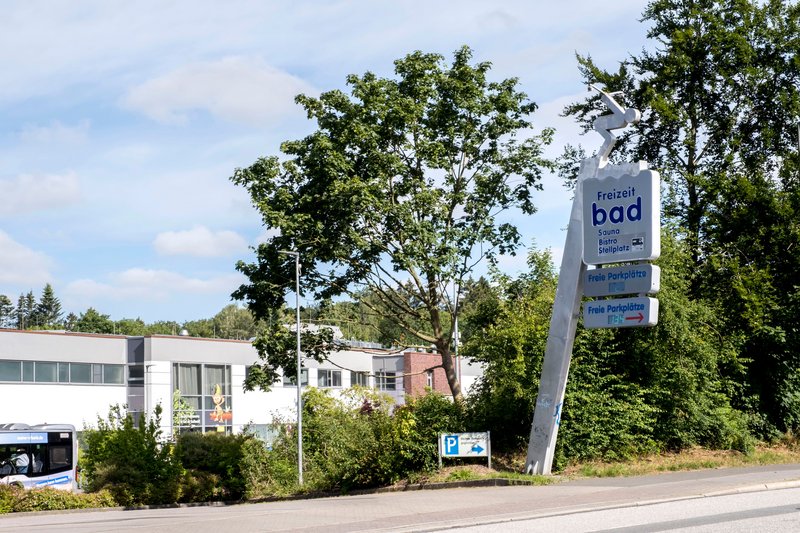 Eingangsschild eines Freizeitbads mit Parkplatzhinweisen, umgeben von Bäumen und einem Gebäude im Hintergrund.