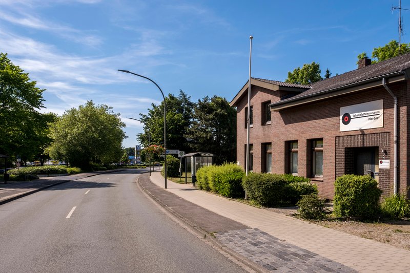 Straße mit dem Deutschen Schallplattenmuseum in Nortorf, umgeben von Bäumen und einem klaren Himmel.