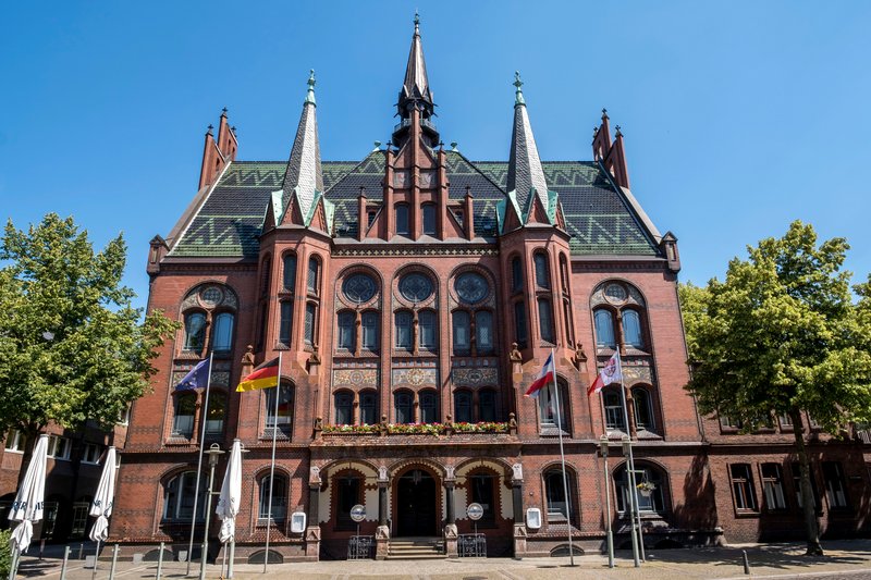 Das Alte Rathaus in Neumünster mit rotem Backstein und Türmen, umgeben von Bäumen und Flaggen.