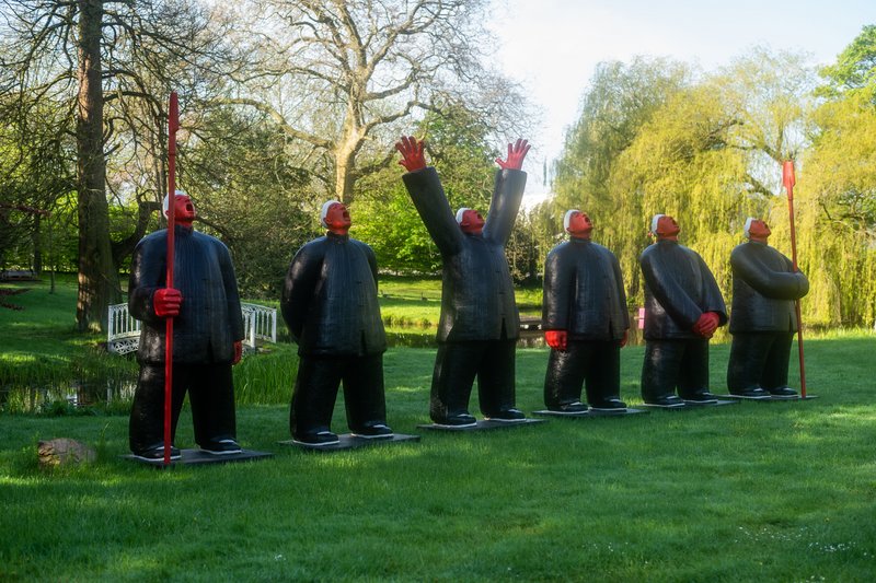 Sechs schwarze Skulpturen mit roten Gesichtern und Händen stehen auf einer Wiese, umgeben von Bäumen.