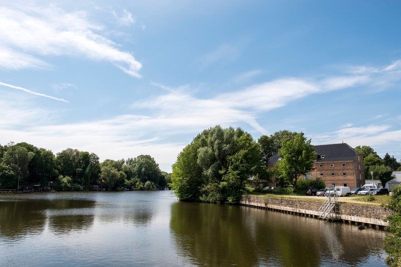 Flusslandschaft mit Bäumen und einem Backsteingebäude am Ufer unter blauem Himmel.