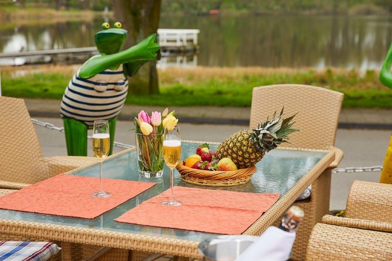 Ein gedeckter Tisch im Freien mit Sektgläsern, einem Obstkorb und Tulpen. Im Hintergrund ist ein See und eine Froschfigur zu sehen.