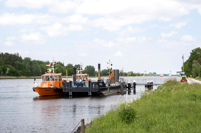 Zwei orangefarbene Boote und ein schwarzes Schiff am Ufer eines Kanals, umgeben von grüner Landschaft und blauem Himmel.