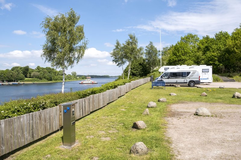 Wohnmobil auf einem Stellplatz am Wasser mit Bäumen und einem vorbeifahrenden Schiff im Hintergrund.