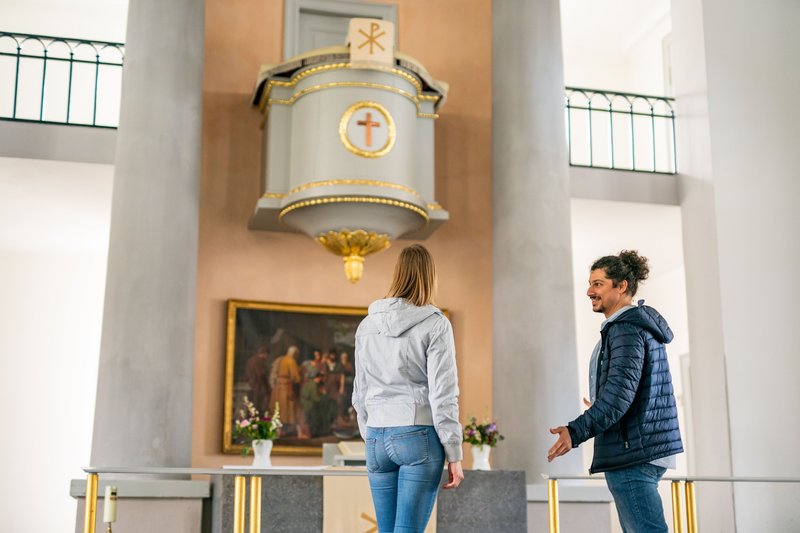 Zwei Personen stehen in einer Kirche vor einer Kanzel und einem Gemälde.