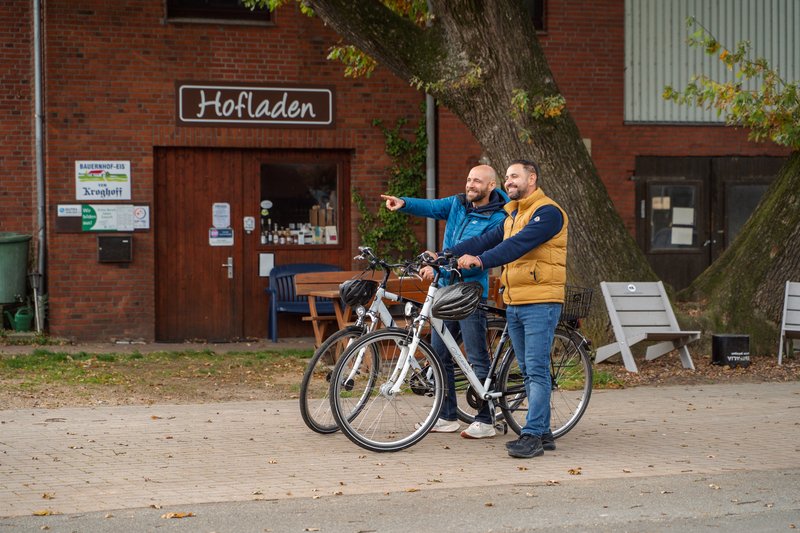 Zwei Männer stehen mit Fahrrädern vor einem Hofladen und zeigen in die Ferne.
