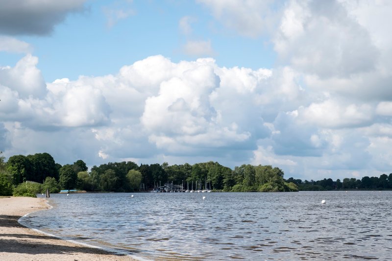 Ein See mit Sandstrand, Bäumen im Hintergrund und Segelbooten am Ufer unter einem bewölkten Himmel.