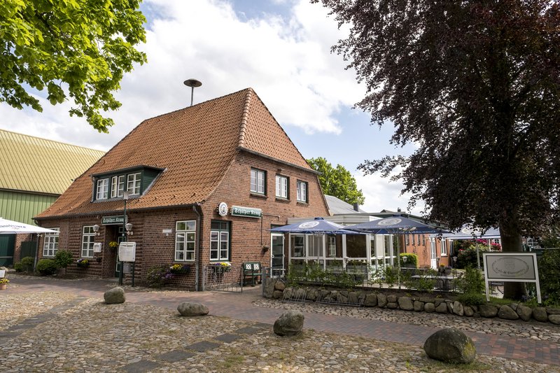 Ein traditionelles Backsteingebäude mit einem roten Ziegeldach, bekannt als Schülper Kroog, umgeben von Bäumen und einem gepflasterten Vorplatz.