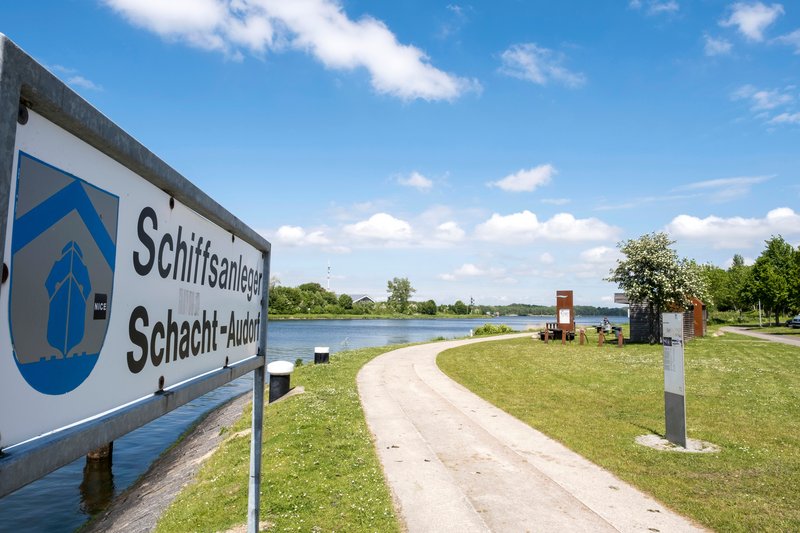 Ein Schild mit der Aufschrift 'Schiffsanleger Schacht-Audorf' steht am Ufer eines Flusses. Ein Weg führt entlang des Wassers, umgeben von grüner Landschaft.