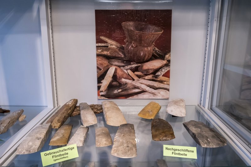 Ausstellung von Flintbeilen in einer Vitrine mit einem Bild von Keramik im Hintergrund.