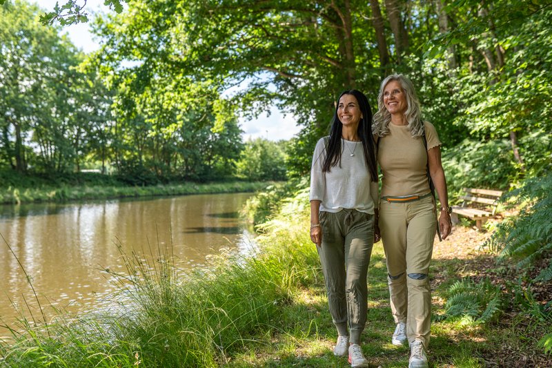 Zwei Frauen spazieren auf einem Waldweg entlang eines Flusses, umgeben von grünen Bäumen und Gras.