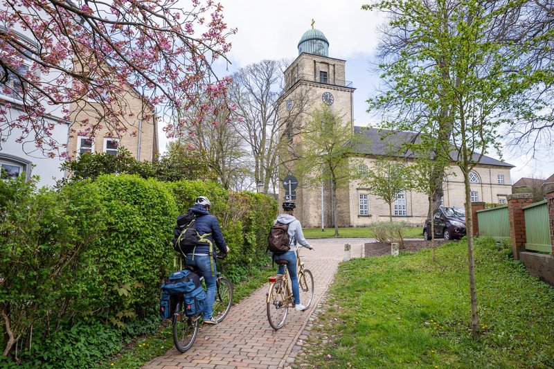 Zwei Radfahrer fahren auf einem Weg zur Vicelinkirche in Neumünster, umgeben von blühenden Bäumen und grüner Vegetation.
