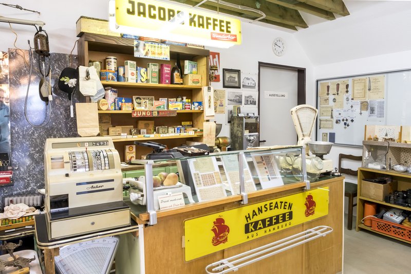 Ein historischer Laden mit alter Registrierkasse, Waage und Regalen voller Vintage-Produkte. Ein Schild mit 'Jacobs Kaffee' hängt oben.