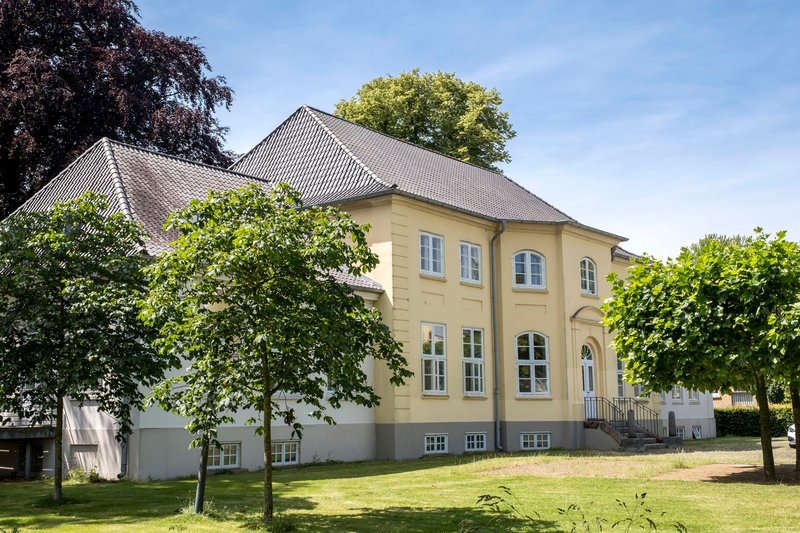 Historisches Gebäude mit gelber Fassade und grauem Dach, umgeben von Bäumen und Rasen.