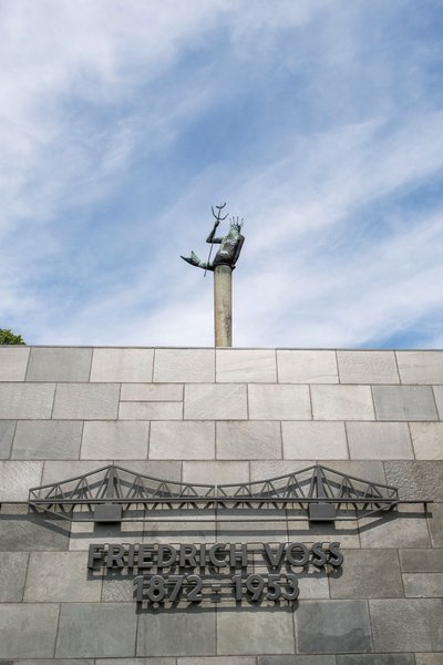 Statue von Neptun auf einer Säule über einer Mauer mit der Inschrift 'Friedrich Voss 1872-1953'.