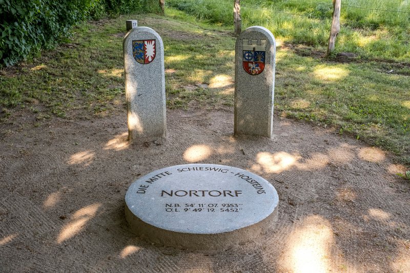 Steinmarkierung des geografischen Mittelpunkts von Schleswig-Holstein in Nortorf, umgeben von zwei Steinsäulen mit Wappen.