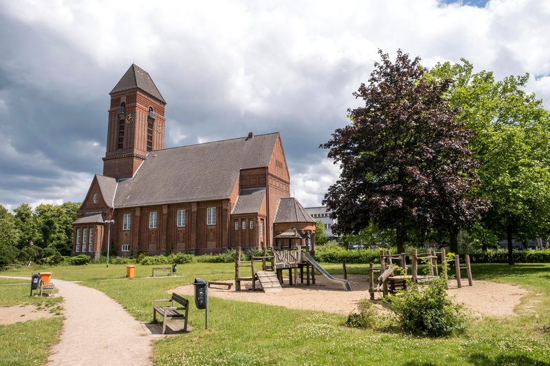 Die Anscharkirche mit einem angrenzenden Spielplatz im Anscharpark, umgeben von Bäumen und einem bewölkten Himmel.