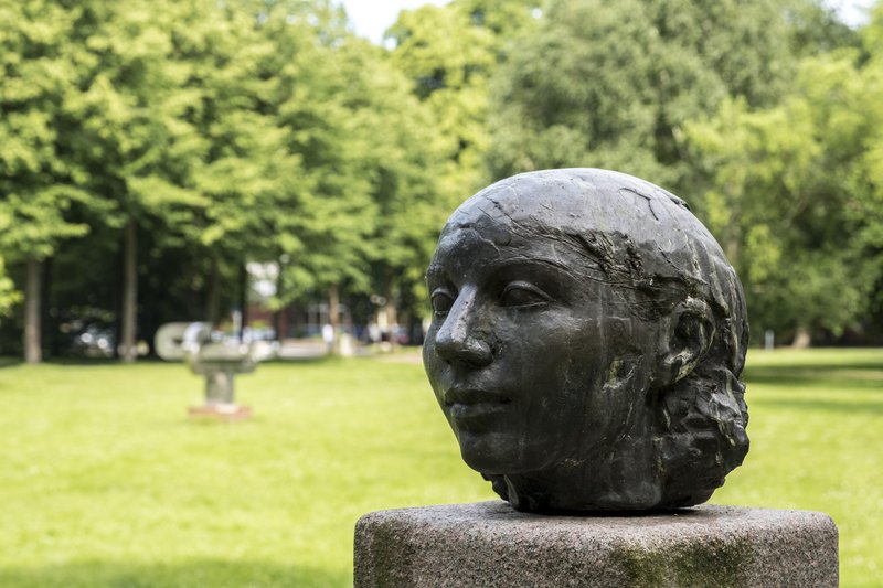 Bronzeskulptur eines Kopfes auf einem Sockel im Stadtpark, umgeben von grünen Bäumen.