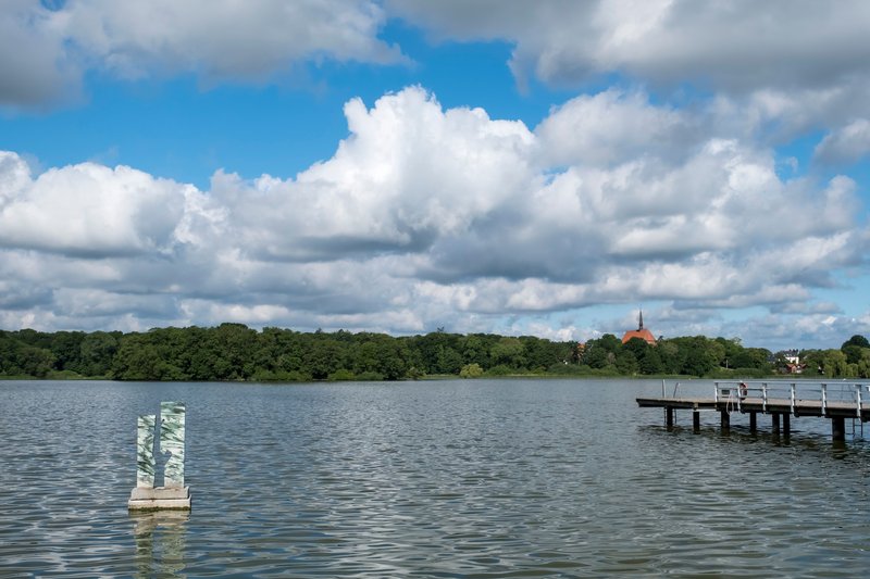 Ein See mit einem Steg und einer Skulptur im Wasser, umgeben von Bäumen und einem Kirchturm im Hintergrund unter einem bewölkten Himmel.