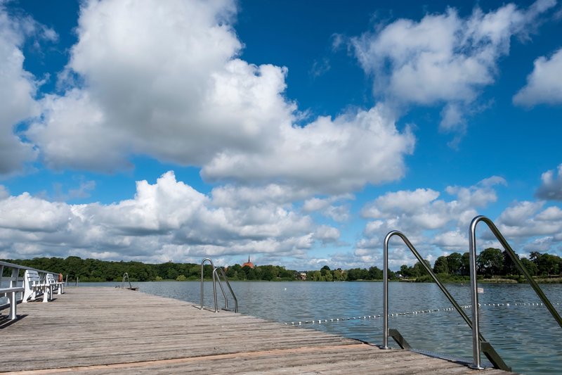 Ein hölzerner Badesteg führt in den Bordesholmer See unter einem blauen Himmel mit weißen Wolken.