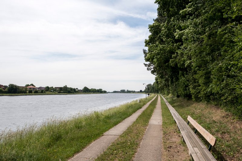 Lange Holzbank entlang eines Kanals mit Bäumen und einem Gehweg.
