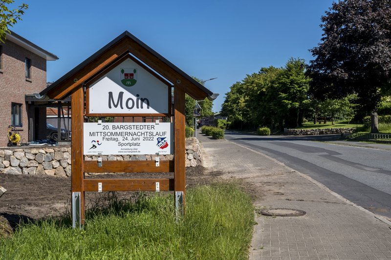 Ein Schild mit der Aufschrift 'Moin' und Informationen zum Bargstedter Mittsommernachtslauf steht am Straßenrand.