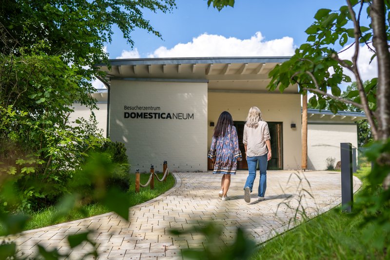 Zwei Personen gehen auf das Besucherzentrum Domestica Neum im Arche Warder zu, umgeben von grünen Bäumen und einem gepflasterten Weg.