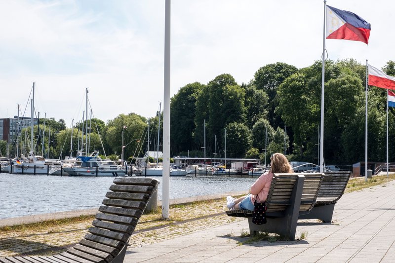 Eine Person sitzt auf einer Bank am Obereiderhafen in Rendsburg, mit Blick auf Segelboote und Flaggen im Hintergrund.