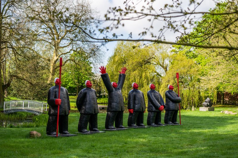 Eine Reihe von Skulpturen mit roten Gesichtern und Händen in einem grünen Park mit Bäumen und einer Brücke im Hintergrund.